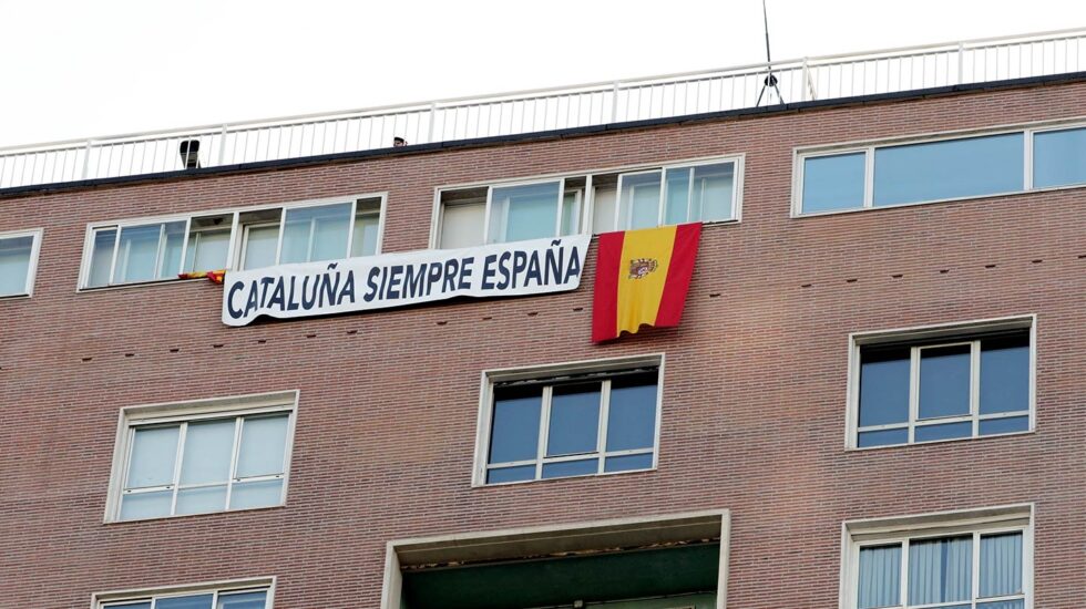 Pancarta junto a la bandera española en uno de los edificios próximos al lugar donde se desarrolla el desfile del Día de la Fiesta Nacional, al que asiste el Gobierno en pleno y la mayoría de líderes políticos.