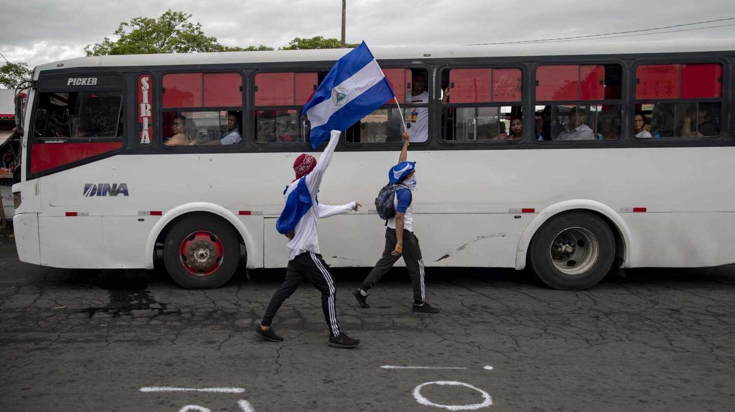 Dos jóvenes con las caras tapadas y una bandera de Nicaragua caminan frente a un vehículo de transporte público durante una protesta por 6 por el ciento del presupuesto nacional a las universidades, y la autonomía en las mismas hoy, jueves 2 de agosto de 2018, en el día numero 107 de protestas contra el gobierno de Daniel Ortega, frente a la Universidad Centroamericana (UCA), en Managua (Nicaragua). Los alumnos de la UCA, quienes habían organizado un plantón para este jueves para reclamar los fondos de ley que el Gobierno había negado al centro de estudios superiores, siguieron adelante con la protesta, pese a que la pasada noche, el Ejecutivo informó de que en el mes de agosto realizará el desembolso correspondiente.