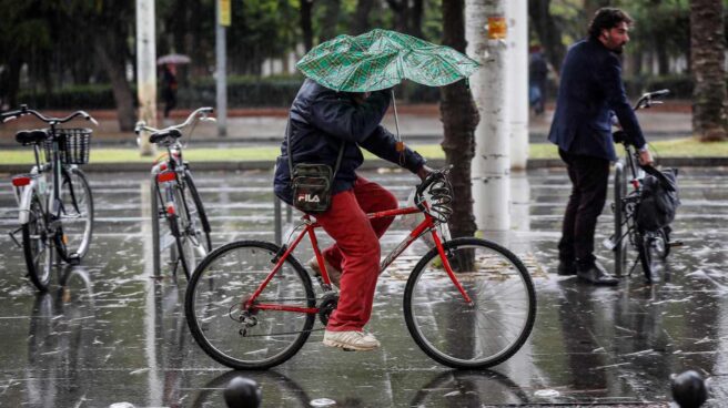 Una persona circula en bicicleta mientras intenta resguardarse de la lluvia con un paraguas, en el centro de Sevilla.