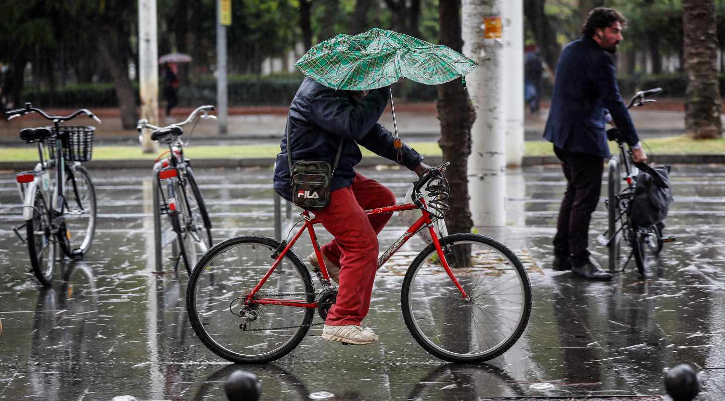 Una persona circula en bicicleta mientras intenta resguardarse de la lluvia con un paraguas, en el centro de Sevilla.
