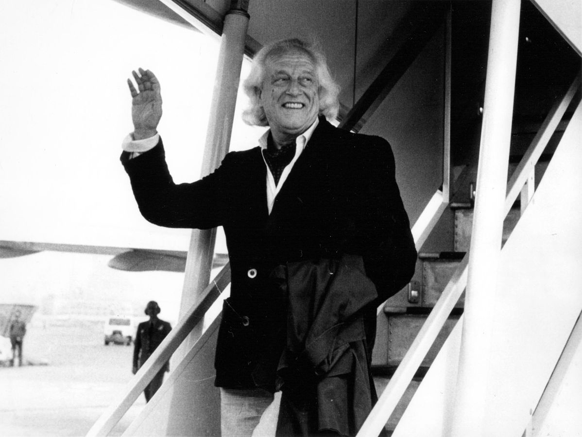 Rafael Alberti, bajando de un avión, saludando con la mano sonriente, llegando a España. Foto en blanco y negro.
