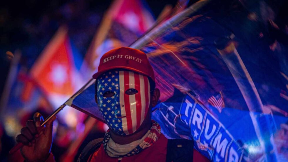 Un simpatizante de Donald Trump viste careta con la bandera estadounidense y una gorra que dice "Mantén (América) grandiosa" mientras espera los resultados de las elecciones presidenciales en Miami