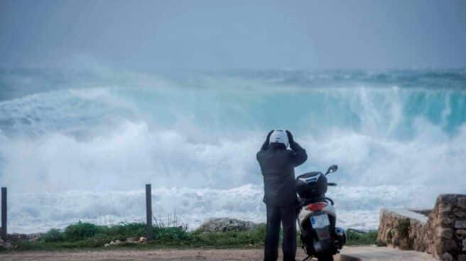 Un motorista se prepara para montar en su scooter mientras grandes olas rompen en Binianolla (Menorca).