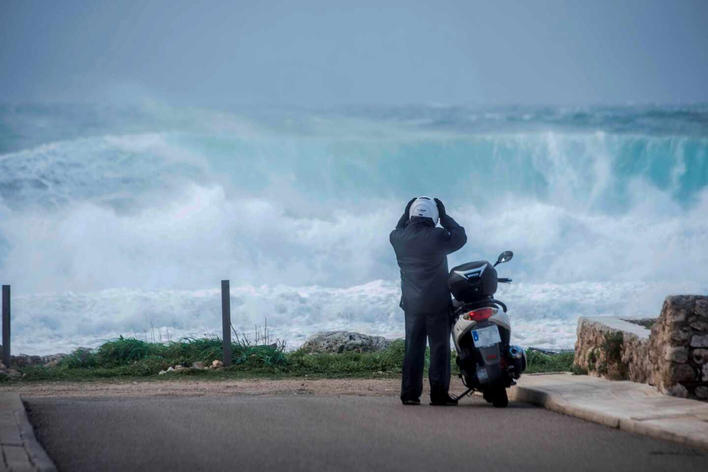 Un motorista se prepara para montar en su scooter mientras grandes olas rompen en Binianolla (Menorca).