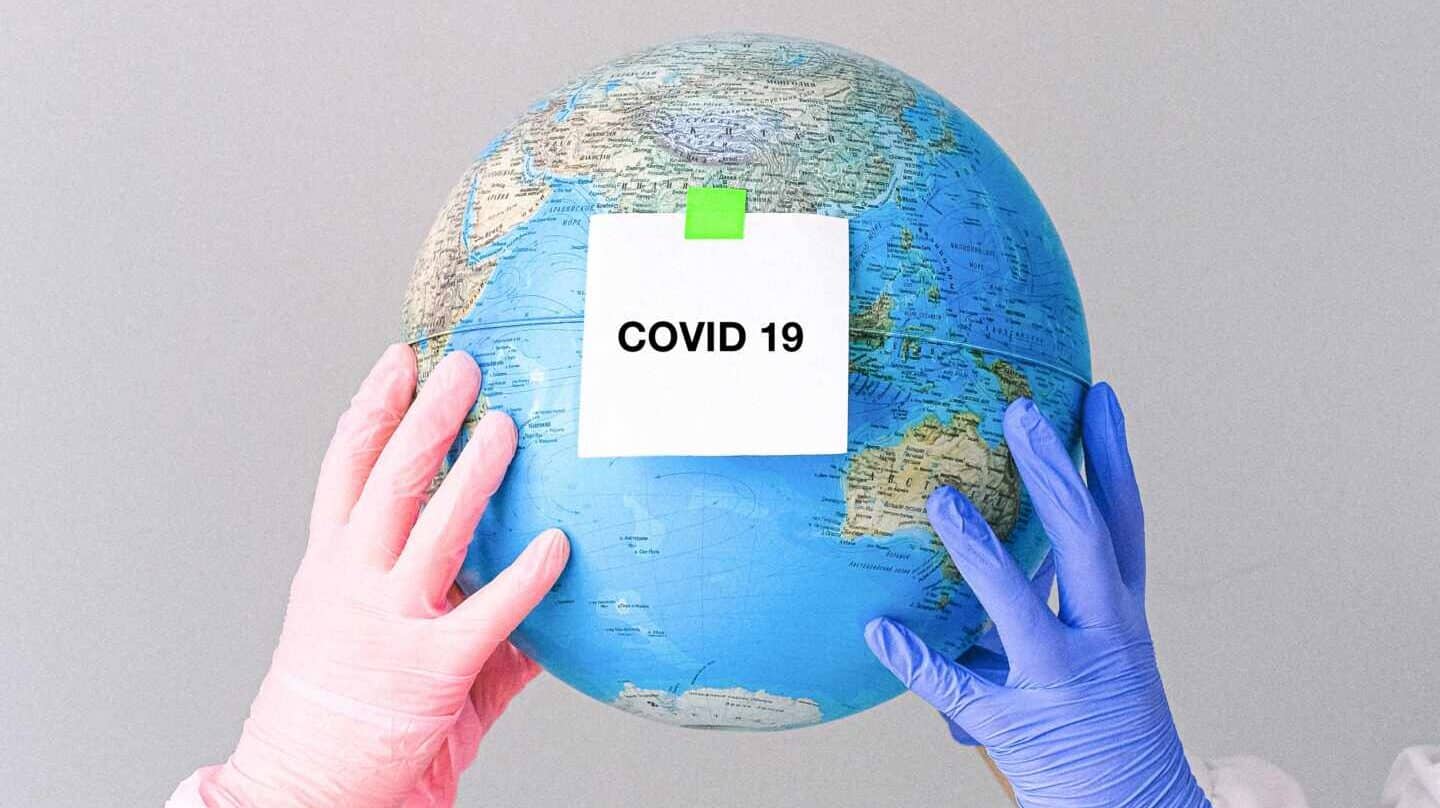 Dos manos con guantes sujetan un globo terráqueo con una nota en la que se lee "Covid-19".