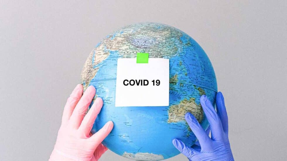 Dos manos con guantes sujetan un globo terráqueo con una nota en la que se lee "Covid-19".