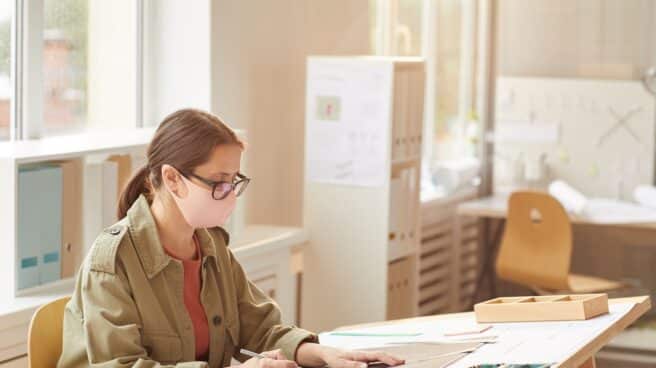 side-view-portrait-of-female-architect-wearing-mask-while-sitting-at-drawing-desk-in-sunlight