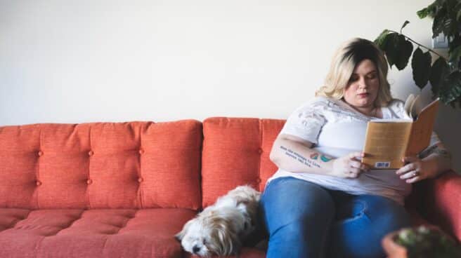 Una persona con obesidad lee un libro sentada en un sofá al lado de un perro.
