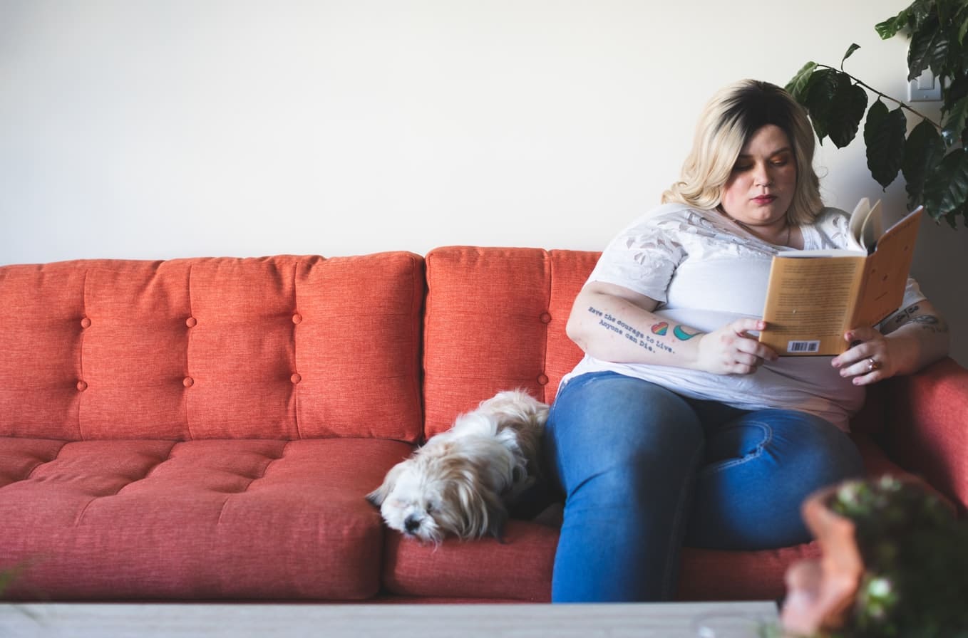 Una persona con obesidad lee un libro sentada en un sofá al lado de un perro.