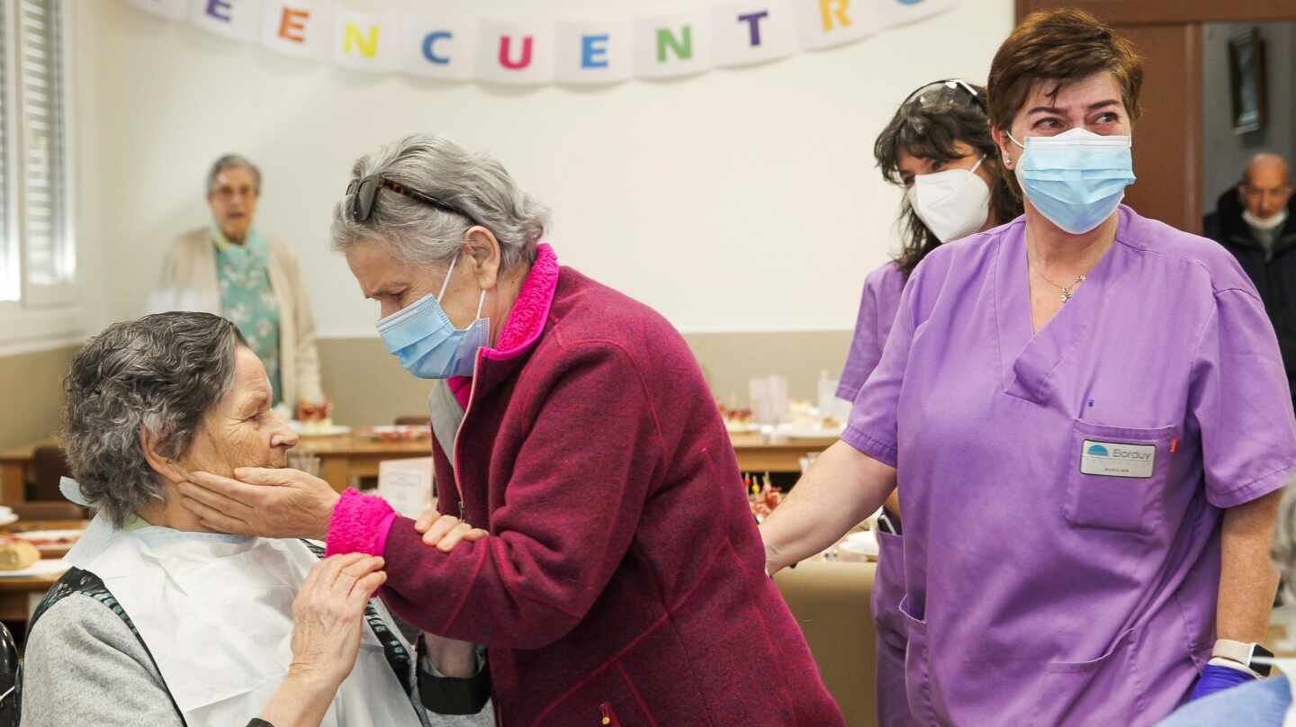 Dos personas mayores reencontrándose en una residencia de ancianos con personal sanitario y mascarillas.