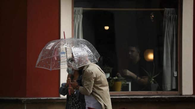 Dos personas pasan por delante de una terraza de un bar protegidos bajo un paraguas.