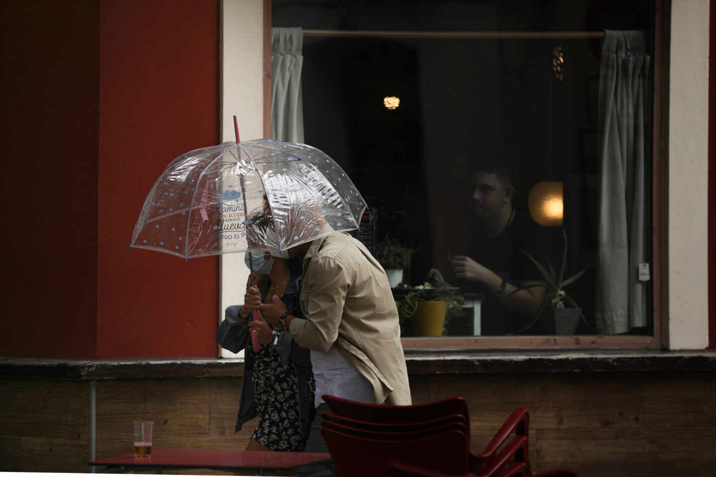Dos personas pasan por delante de una terraza de un bar protegidos bajo un paraguas.