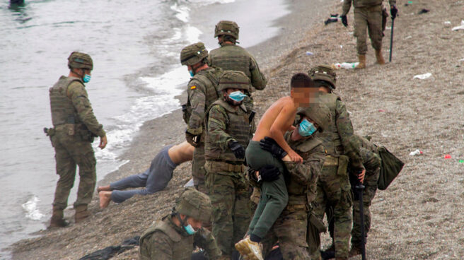 Un miembro del Ejército sostiene en brazos a un joven sin camiseta en la playa del Tarajal