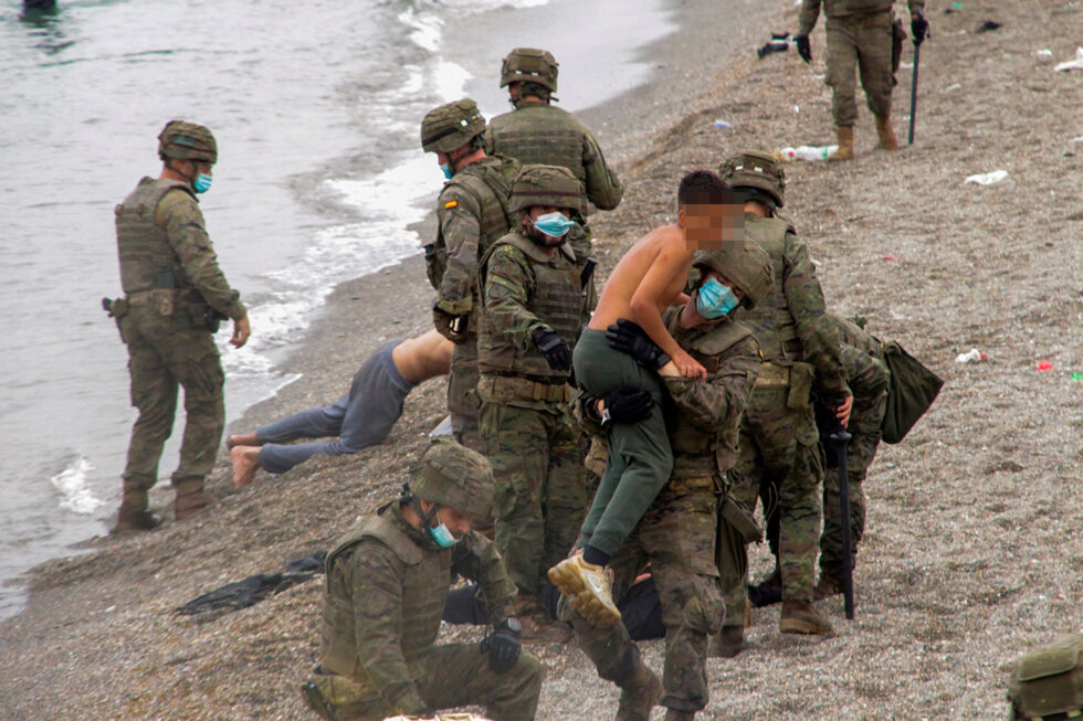 Un miembro del Ejército sostiene en brazos a un joven sin camiseta en la playa del Tarajal