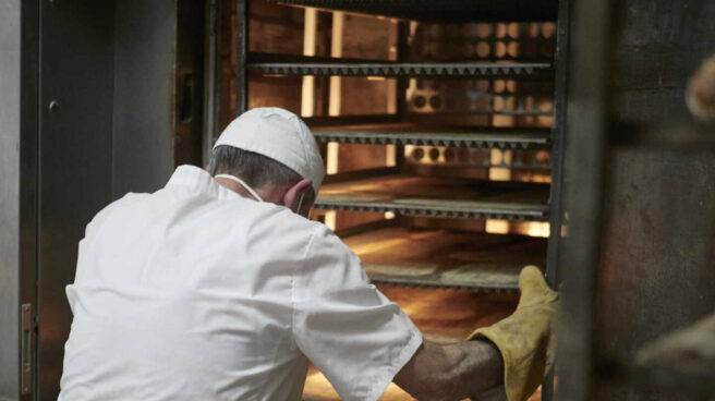 Un trabajador protegido con mascarilla y guantes maneja bandejas para cocer el pan en un obrador.