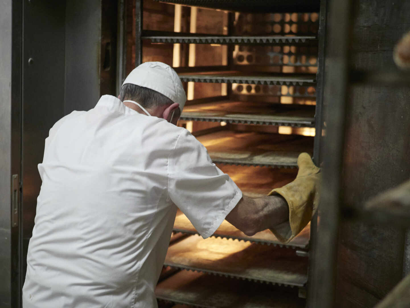 Un trabajador protegido con mascarilla y guantes maneja bandejas para cocer el pan en un obrador.
