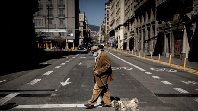 Un hombre con una mascarilla pasea a su perro cruzando una calle de Barcelona.