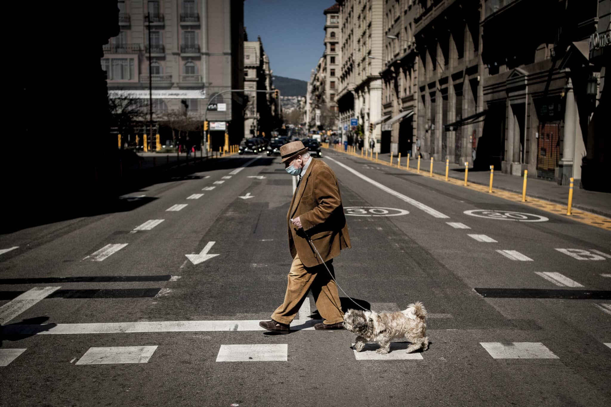 Un hombre con una mascarilla pasea a su perro cruzando una calle de Barcelona.