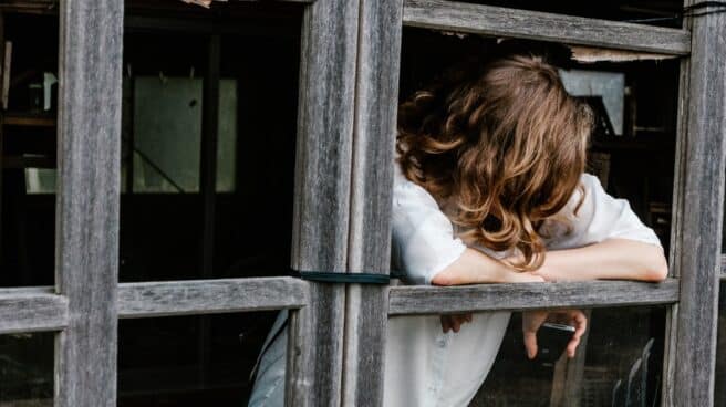 acoso-escolar-vacaciones-continua una joven con el móvil en la mano mira hacia el suelo apoyada en una ventana.