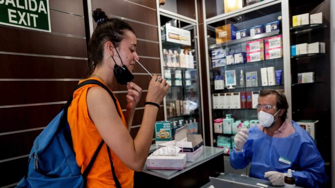 Cuanto cuestan los test de antígenos de las farmacias en España Chica realizándose un test de antígenos de autodiagnóstico en una farmacia junto a un farmacéutico