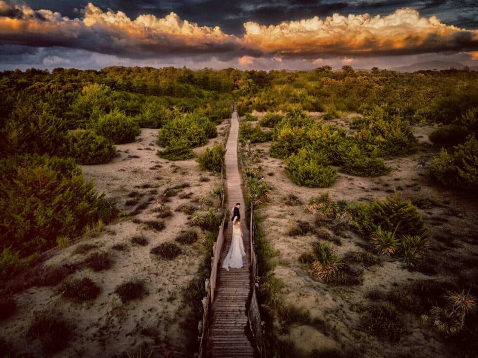 Fotografía vista de dron de una pareja de novios caminando por un camino de madera en medio del campo.