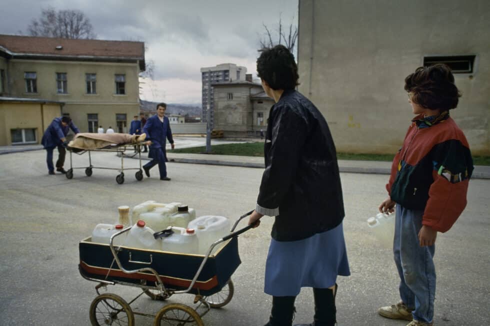 Una madre y su hija cargadas de bidones de agua que han llenado en una fuente pública observan el traslado al depósito de cadáveres de una persona víctima de un bombardeo. La vida, anclada en la más pura supervivencia, se cruza con la muerte en una calle de Sarajevo durante su brutal cerco que duró tres años y medio. Este tipo de escenas eran muy cotidianas y diezmaban la capacidad de resistencia de los ciudadanos que vivían sin agua, luz y calefacción. Los sitiadores, armados con artillería pesada y sólidamente pertrechados en las colinas que rodeaban la capital bosnia y en algunos de sus barrios, sometieron a un infierno diario a los centenares de miles de ciudadanos. Los cementerios crecían cada día por culpa de un espantoso e inútil goteo de muerte. Sarajevo (Bosnia-Herzegovina), setiembre de 1993