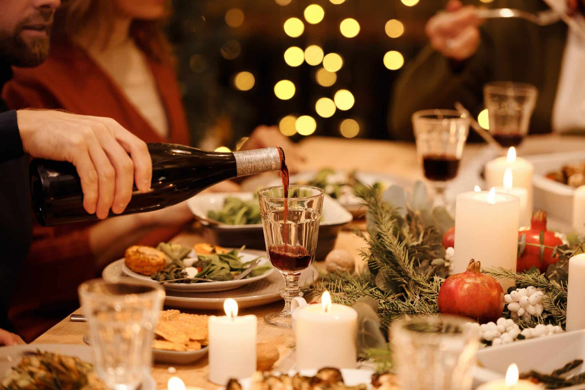 Una mano sirve una copa de vino en una mesa de Navidad, con velas y ramas.