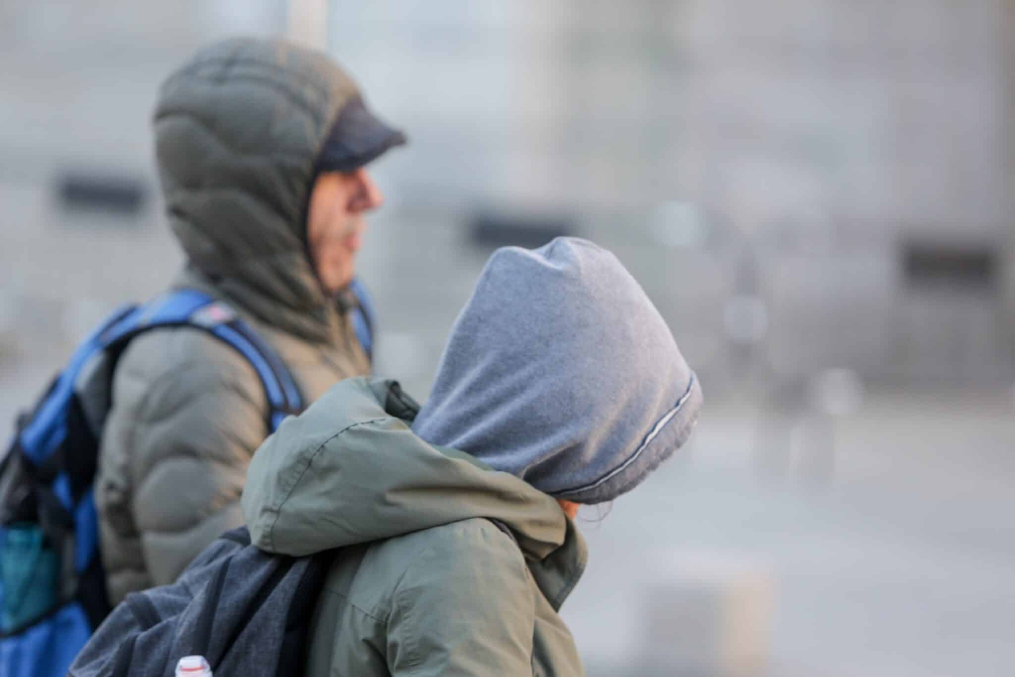 Dos jóvenes se protegen del frío y la lluvia con abrigos y gorros mientras pasean durante un día de viento por Madrid