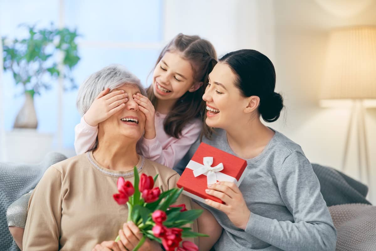 Dos mujeres de diferentes edades y una niña sonrientes entre ramos de flores y regalos
