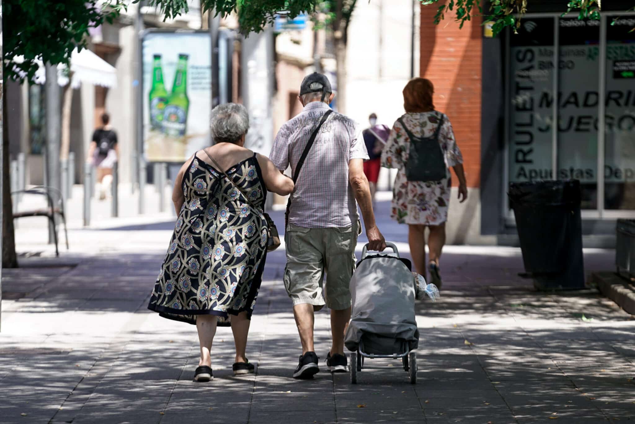 Una pareja de ancianos camina por la calle con un carro de compra.