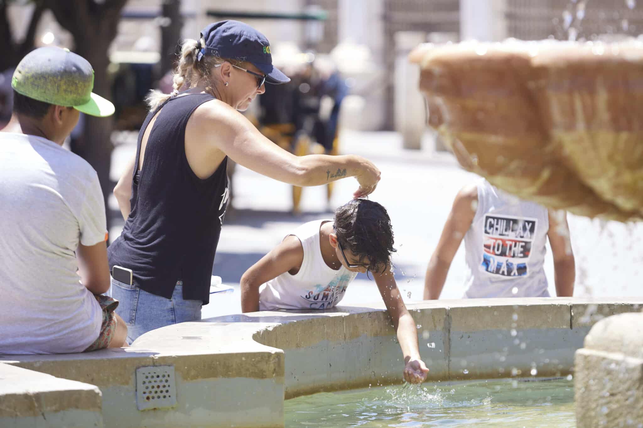 Una madre le echa agua a su hijo, por la cabeza, en la fuente de la plaza Virgen de los Reyes en el primer día de la segunda ola de calor