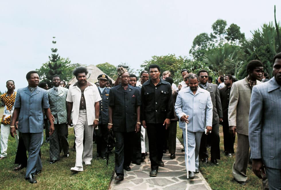 Ali, flanqueado por el promotor Don King y su manager Herbert. Mohamed es recibido por Mobutu Sese Seko, dictador de Zaire, con su característico sombrero de piel de leopardo. Mobutu costeó con 10 millones de dólares el evento sabiendo que el combate llevaría a Zaire a la escena deportiva internacional.