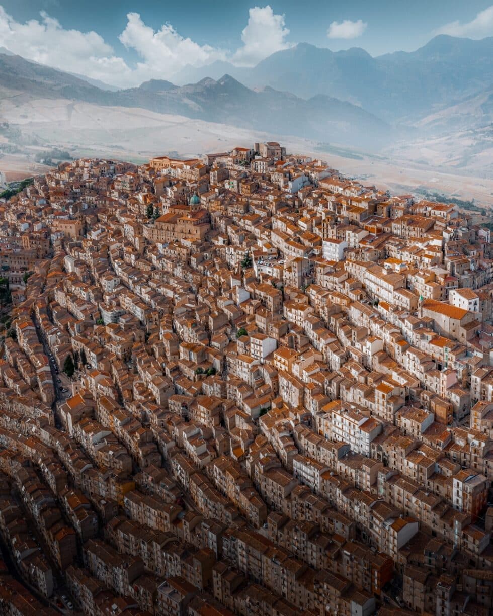 Vista aérea de Gangi, localidad de la campiña siciliana en la provincia de Palermo. La imagen fue tomada en una mañana de principios de verano. Los techos de las casas anaranjadas dan movimiento a la imagen, mientras los picos de las montañas se alzan al fondo.