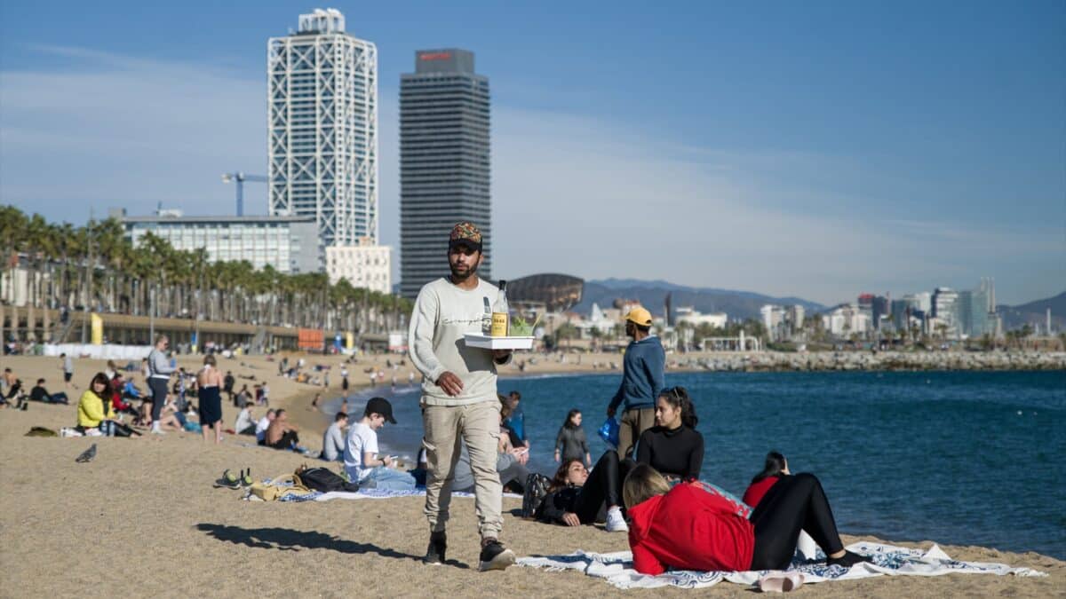 Un camarero lleva bebidas a las personas que disfrutan de las altas temperaturas en la playa de la Barceloneta, en Barcelona.