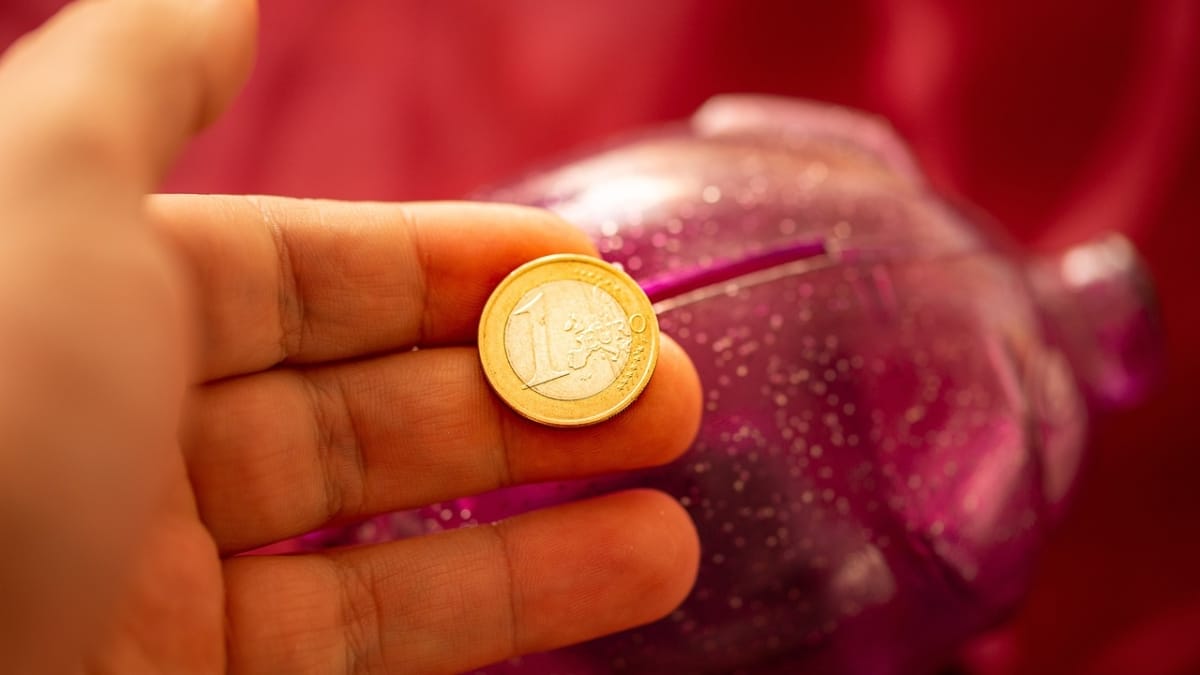 Una mano sostiene una moneda de un euro encima de una hucha.