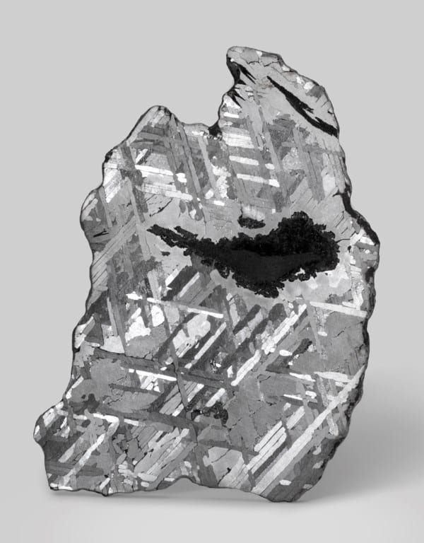 Lámina pulida del meteorito Toluca, un hierro del grupo IAB formado en la corteza de un cuerpo primitivo tras un impacto que fundió metal y silicatos. Cayó a la Tierra hacia el 8000 a. C. cerca de la actual Ciudad de México y fue utilizado por poblaciones indígenas antes de su “descubrimiento” oficial en 1776 durante la colonización española. Su patrón grabado —muy apreciado por su brillo y geometría— ha sido usado incluso por la firma relojera suiza Corum en esferas de relojes fabricadas con láminas finas de Toluca. Esta pieza conserva la sección completa, con una gran inclusión de grafito rodeada de troilita.