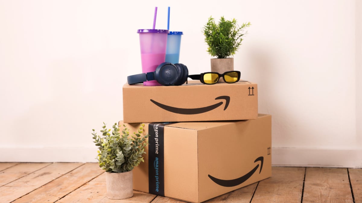 Dos cajas de cartón de Amazon apiladas sobre un suelo de madera, con cinta de Amazon Prime, unos auriculares de diadema, gafas de sol amarillas, dos vasos de colores con pajita y plantas en maceta