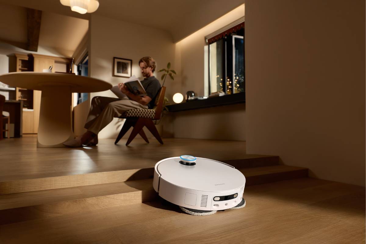 Robot aspirador blanco con anillo azul sobre suelo de madera; al fondo, un hombre lee sentado en una silla.