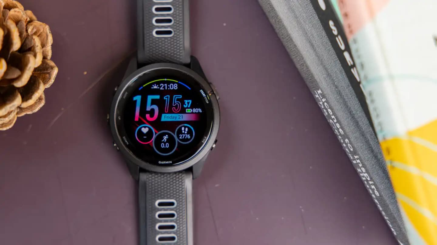 Reloj Garmin Forerunner 265 con correa gris oscura y esfera AMOLED encendida sobre una mesa morada, junto a una piña y un libro