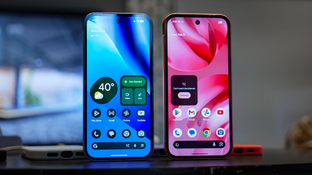 Dos móviles Google Pixel de pie, uno en azul lila y otro en rosa coral, con la pantalla de inicio encendida sobre una mesa oscura