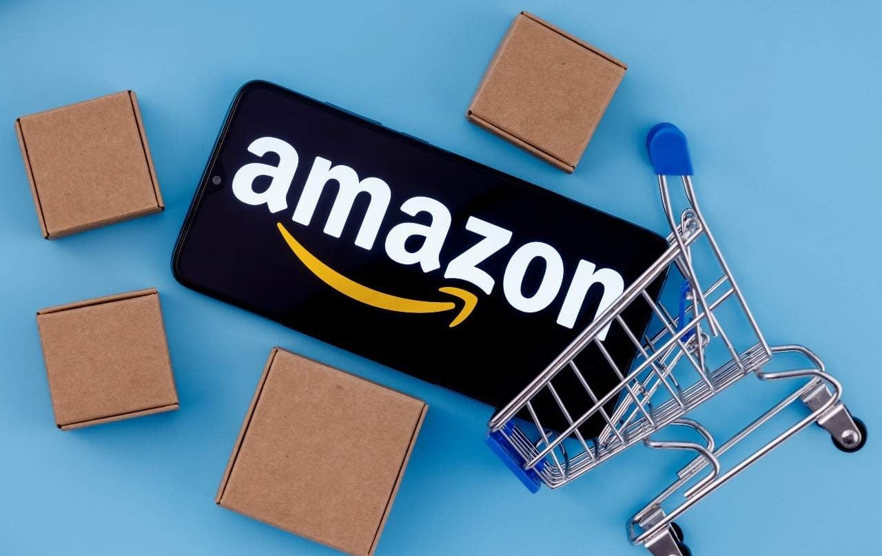 Teléfono con logotipo de Amazon, rodeado de cajas de cartón y un carrito de compra volcado sobre fondo azul.