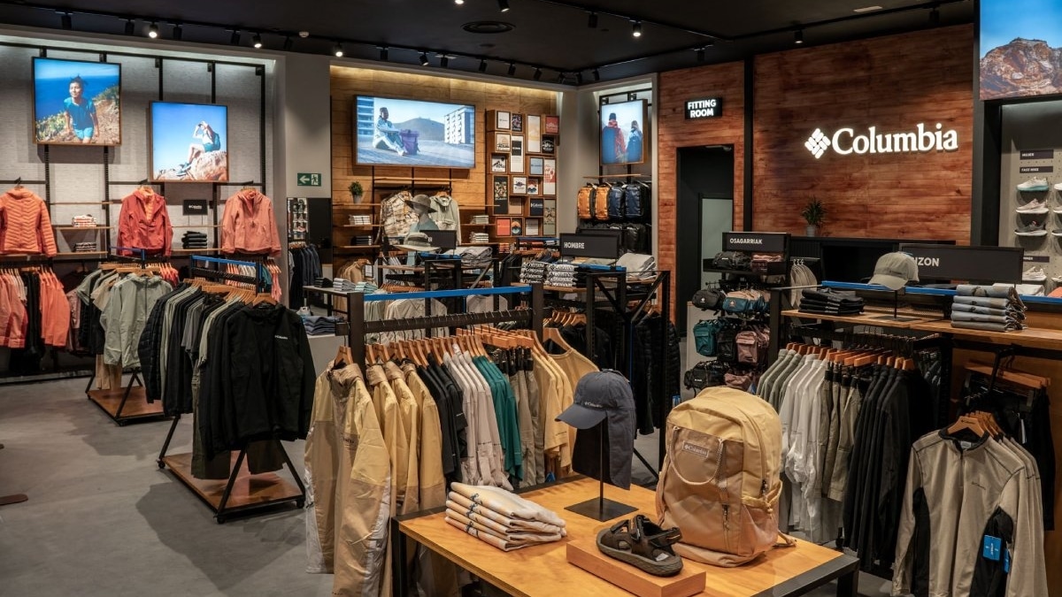 Interior de una tienda Columbia con chaquetas, forros polares y camisas colgadas en percheros, una mesa con una mochila, una gorra y unas sandalias, y el logotipo de Columbia en una pared de madera