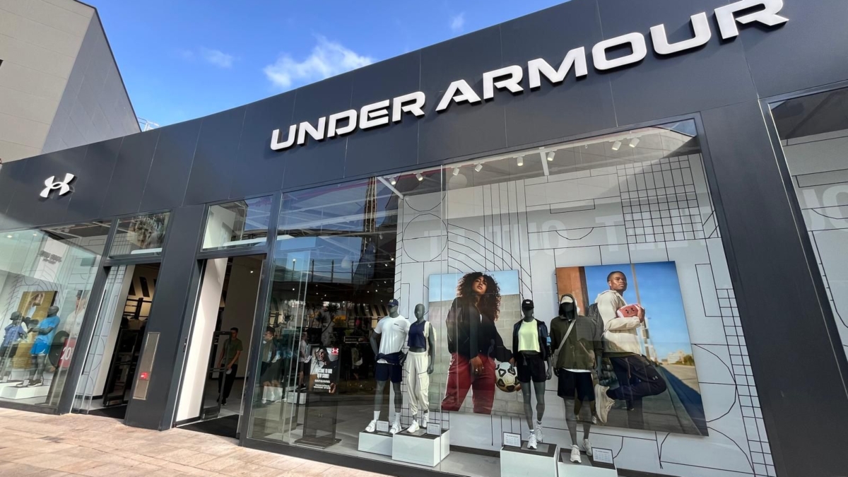 Fachada negra de tienda Under Armour con maniquíes deportivos en el escaparate bajo un cielo azul.